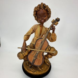 Könnte beinhalten: Vintage Figur einer Person, die Cello spielt. Die Figur trägt ein goldfarbenes Samtkostüm und hat einen dunklen Teint. Das Cello ist braun mit einem goldenen Bogen. Der Sockel ist gold und schwarz.