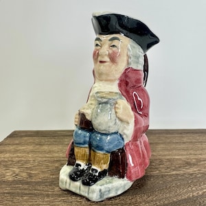 Puede incluir: Una jarra de cerámica con forma de hombre que lleva un abrigo rojo, un sombrero negro y pantalones azules. Sostiene una jarra más pequeña en su mano derecha.