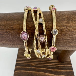 May include: A set of three gold-tone metal bangles featuring a hammered texture. The bracelets are adorned with various round and oval-shaped faceted crystals in shades of clear, light pink, and dark pink, set securely into the metal bands.