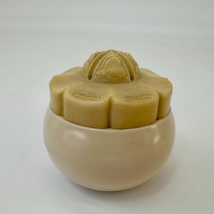Puede incluir: Un tarro vintage de color crema con tapa en forma de flor. La tapa presenta una abeja tallada y la palabra "Yardley London". El tarro tiene una base redondeada y un color crema a juego. La tapa es de color marrón claro.