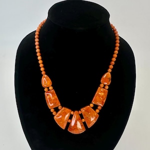 May include: Orange beaded necklace with a series of geometric, orange-toned pendants. The necklace is displayed on a black mannequin. The beads and pendants have a marbled appearance.