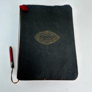 Puede incluir: Un libro antiguo encuadernado en cuero negro con letras doradas y un lápiz rojo y plateado sujeto con una cuerda. El libro tiene un emblema dorado en la portada. El lápiz es rojo con punta plateada.