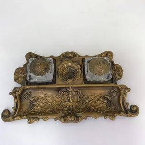 May include: A gold-toned metal inkstand featuring two square glass inkwells with matching ornate metal lids. The base has an intricate scrollwork design and a central shell-shaped indentation for a pen, with a wide tray for writing implements.
