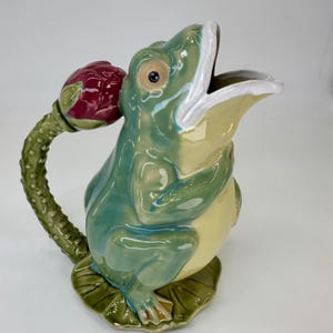 Op de afbeelding: Een keramische kikkerkan in groen en geel, met een open mond. De handgreep is een groene stengel met een rode bloemknop. De kan staat op een groene leliebladbasis. Een decoratief item.