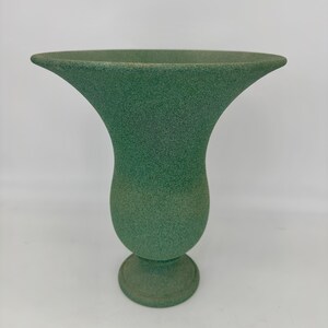 May include: A green textured vase with a wide, flared top and a rounded base. The vase has a speckled appearance and a pedestal base. The vase is a muted green color and is likely made of ceramic or a similar material.