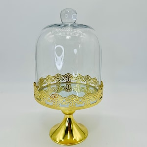 May include: A clear glass cloche with a rounded top and a small knob. The cloche sits on a gold-colored base with a decorative, openwork design around the edge. The base has a pedestal shape.