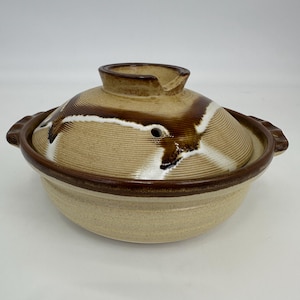 Puede incluir: Una olla donabe japonesa de cerámica beige con tapa. La tapa tiene detalles en marrón y blanco y una pequeña abertura. La olla tiene un borde y asas marrones. La olla está diseñada para cocinar y servir.