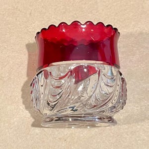 May include: A clear glass container with a ruby red rim. The container has a wavy, textured design on the lower portion and a scalloped edge on the red rim. The container is likely a decorative piece or a small serving dish.