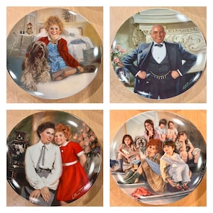 May include: Four decorative plates with scenes from the musical "Annie." The plates show characters and scenes, including Annie with a dog, a man in a suit, and group portraits. Each plate has a glossy finish.