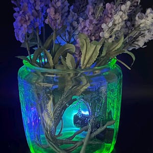 May include: A clear glass jar with a green and blue glow, filled with purple and white flowers. The jar has a textured design and is set against a dark background, creating a vibrant and eye-catching display.