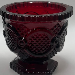 Puede incluir: Un cuenco de pedestal de vidrio rojo rubí con un patrón texturizado detallado. El cuenco presenta un diseño de diamantes y óvalos, con un borde festoneado y una base cuadrada. El cuenco mide aproximadamente 10 cm de alto.