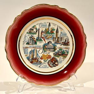 Puede incluir: Un plato de cerámica rojo y blanco con un borde dorado. El plato presenta una ilustración colorida de monumentos de Ohio, incluyendo el Capitolio del Estado, el Salón de la Fama del Rock and Roll y el Salón de la Fama del Fútbol Profesional. El plato está inscrito con la palabra "Ohio" en letras doradas.