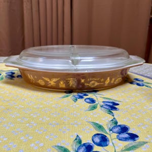 Puede incluir: Una fuente para horno Pyrex de color ámbar con tapa de cristal transparente. El plato presenta un patrón decorativo de símbolos dorados alrededor del borde. El plato es de forma ovalada y está sobre un mantel amarillo con un estampado floral azul.