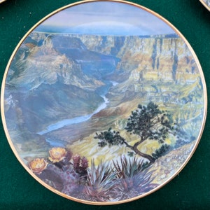 Op de afbeelding: Een decoratieve plaat met een gouden rand met een landschap van de Grand Canyon. Het kunstwerk toont de gelaagde rotsformaties van de canyon, een rivier en woestijnplanten, waaronder cactussen en een kleine boom. De plaat staat tegen een groene achtergrond.