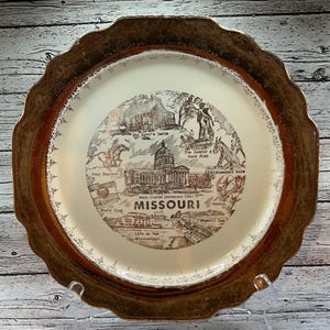 May include: A vintage decorative plate with a gold rim and a cream-coloured centre. The centre features a sepia-toned illustration of Missouri landmarks, including the State Capitol and the words "Missouri" and "State Capital (Jefferson City)".