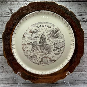 Peut inclure: Une assiette décorative avec un bord doré et un centre crème représentant une illustration détaillée des monuments du Kansas, dont le Capitole de l'État et Mushroom Rock. L'assiette a un bord festonné et est exposée sur un support transparent.