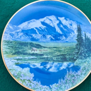 Op de afbeelding: Een blauw en wit porseleinen bord met een schilderachtig beeld van een besneeuwde bergketen, een meer en een bos. Het bord heeft een gouden rand en een afbeelding van beren op de voorgrond.