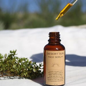 May include: A brown glass bottle of Desert Sun Face Serum with a dropper dispensing the serum. The label lists ingredients like creosote, golden jojoba oil, and rosehip oil. The bottle is 1 fl. oz.