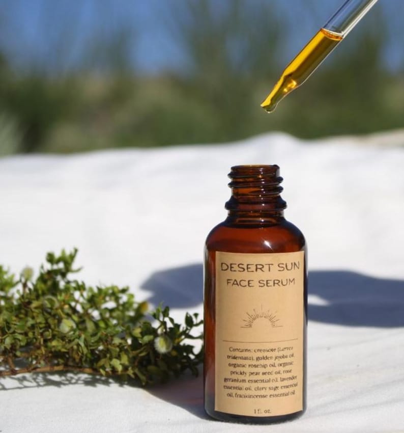 May include: A brown glass bottle of Desert Sun Face Serum with a dropper dispensing the serum. The label lists ingredients like creosote, golden jojoba oil, and rosehip oil. The bottle is 1 fl. oz.
