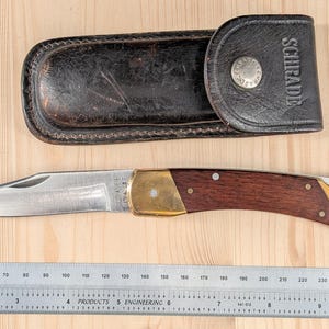 May include: A Schrade folding pocket knife with a silver steel blade, brass bolsters, and dark wood handle scales, positioned above its dark brown leather sheath embossed with the Schrade logo, alongside a metal ruler measuring up to 10 inches.