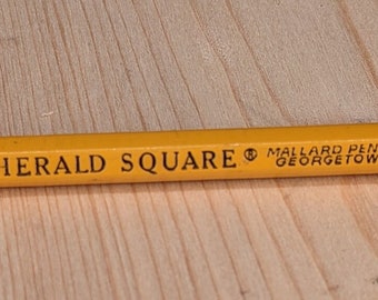 May include: A yellow pencil with the text "SUPER HERALD SQUARE" and "MELLARD PENCIL CO. - 222 -" printed on the side. The pencil has a pink eraser and a silver metal band. The pencil is lying on a light brown wooden surface.