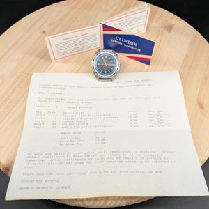May include: A vintage Clinton watch with a black dial and blue bezel, displayed with a repair invoice and warranty cards. The watch has a stainless steel case and a day-date function. The invoice details the watch's repair.