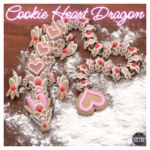 May include: A pink and brown gingerbread dragon with heart-shaped details. The dragon is decorated with pink icing and is lying on a surface covered in white powder.