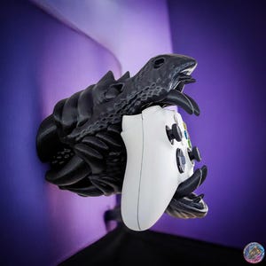 May include: A black dragon-shaped wall mount with a white video game controller in its mouth. The dragon is detailed with scales and has a textured surface.