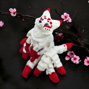 May include: White and red fox figurine with a detailed face and multiple tails. The figurine is made of a soft, textured material and has a whimsical, fairytale-like appearance.