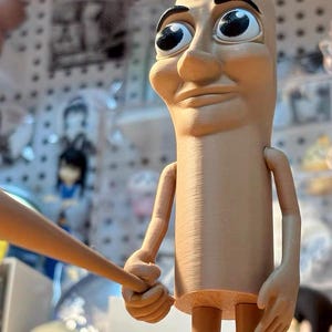 May include: A tan-colored figurine with large eyes and a serious expression. The figurine is holding a wooden baseball bat in its right hand. The figurine is standing on a wooden surface.