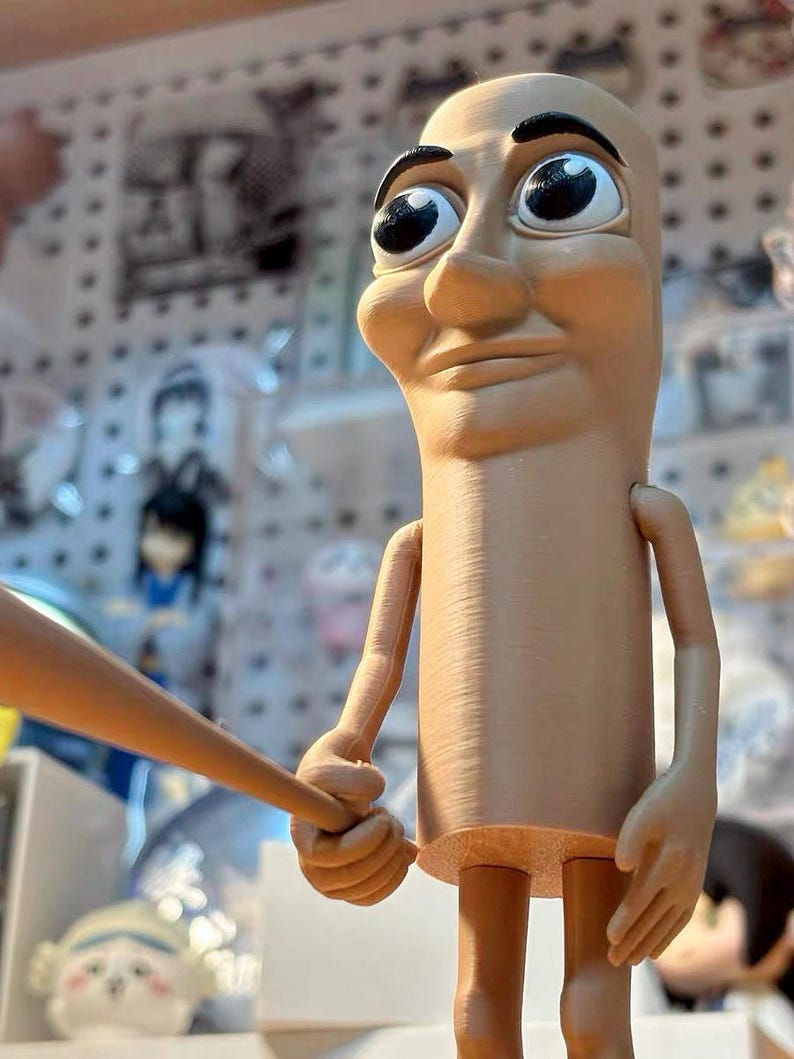 May include: A tan-colored figurine with large eyes and a serious expression. The figurine is holding a wooden baseball bat in its right hand. The figurine is standing on a wooden surface.