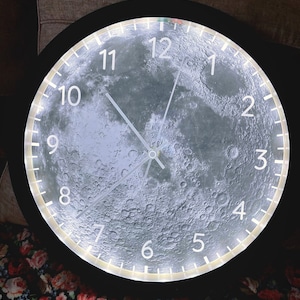 May include: A black and white moon-shaped clock with a black frame and white numbers. The clock face is a detailed image of the moon's surface.
