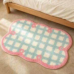 May include: A cloud-shaped rug with a pink border and a checkered pattern in light blue and cream. The rug is placed on a neutral-colored carpet, near a wooden bed frame and white bedding.
