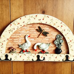 May include: A decorative wall-mounted coat rack with a semi-circular design. It features a brick-patterned background, adorned with goose figurines, a dragonfly, and floral accents. The rack includes three black hooks for hanging items.
