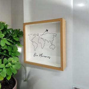 Puede incluir: Una caja de sombra de madera con un fondo blanco que presenta un dibujo de línea negra del mundo con un pequeño avión volando por encima. El texto "Our Memories" está escrito debajo del mapa del mundo.