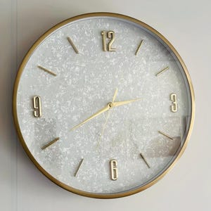 May include: A gold-rimmed clock with a white face and black hour and minute hands. The clock face has a white, sparkly, textured background. The numbers 12, 9, 3, and 6 are in gold.
