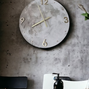 May include: A round, gray concrete clock with gold-colored hands and numbers. The clock is hanging on a gray wall.