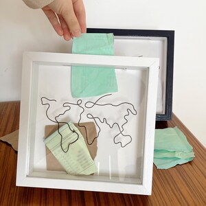 May include: A white shadow box frame with a wire sculpture of the world map inside. The frame is on a wooden surface and there are pieces of green and brown paper inside the frame.