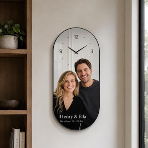 May include: An oval wall clock with a black frame featuring a mirrored surface printed with a photograph of a couple. The clock face has black hands and the numbers 3, 9, and 12. Below the photo, the text reads Henry & Ella October 15, 2026.