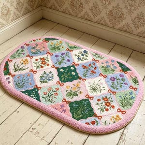 Puede incluir: Una alfombra ovalada con un diseño de patchwork que presenta patrones florales en varios colores. La alfombra tiene un borde rosa y está colocada sobre un suelo de madera. Lazos rojos conectan los cuadrados florales.