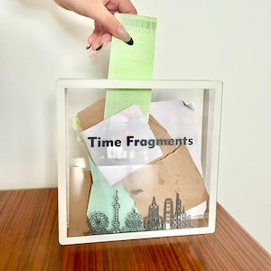 May include: A clear acrylic box with a white frame and the text "Time Fragments" printed on the front. The box is filled with various pieces of paper and a receipt. The box is sitting on a wooden surface.