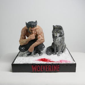 Puede incluir: Una figura esculpida de un hombre musculoso con cabello oscuro y patillas agachado junto a un lobo gris sobre una base nevada. La base presenta una mancha de color rojo que recuerda a la sangre y la palabra WOLVERINE en letras rojas.