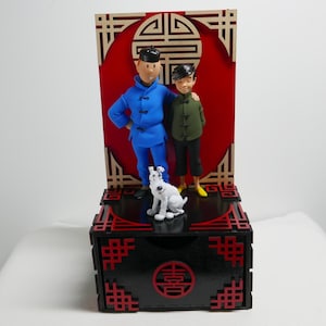 Op de afbeelding: Een decoratieve display met twee figuren en een witte hond op een zwarte doos met rode accenten. De figuren dragen blauwe en groene jasjes in traditionele stijl. Een rode achtergrond met een beige geometrisch frame staat achter de figuren.