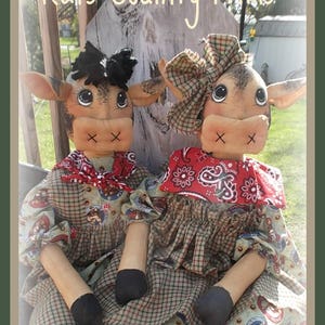 Könnte beinhalten: Zwei handgefertigte Kuhpuppen, die in karierten Kleidern mit roten Bandanas und schwarzen Schuhen gekleidet sind. Die Puppen sitzen auf einer Holzbank und der Text "Bessie & Bertis" steht darunter.