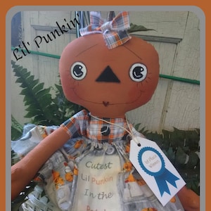 Pode incluir: Boneca de abóbora artesanal com laço e vestido xadrez. A boneca tem um rosto sorridente e usa um vestido com estampa de abóbora. A etiqueta diz "Cutest Lil Punkin In the Patch" e "1st Place Winner". Os textos "Lil' Punkin" e "Kats Country Prims" também são visíveis.