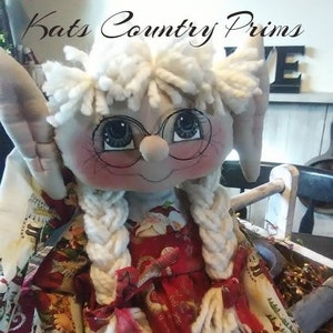 May include: A handmade Christmas elf doll with white yarn hair, a red dress with gold ornaments, and a pair of glasses. The doll is sitting on a wooden chair with a white fabric background.