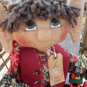 May include: A handmade elf doll with a red and green plaid vest, a red hat, and a green and white plaid shirt. The elf is holding a toy train with a red and white caboose.