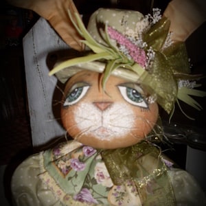 May include: A handmade bunny doll wearing a green and white hat with a floral design and a green and gold ribbon around its neck. The doll has a painted face with brown eyes and a white nose.