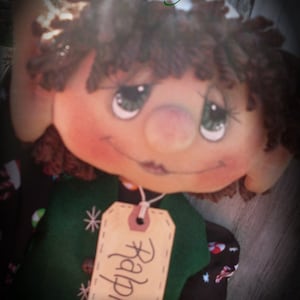 May include: A handmade elf doll with brown yarn hair, wearing a green and black vest with a candy cane pattern. The doll has a tag that says "Ralph" on it.