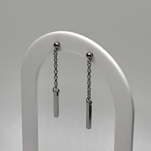 Puede incluir: Un par de pendientes de barra plateados colgando de delicadas cadenas. Los pendientes se exhiben en un soporte blanco en forma de arco con una base de madera, sobre un fondo liso. Diseño sencillo y moderno.